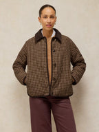 A model wearing a brown and beige plaid quilted jacket with a dark brown corduroy collar, paired with dark plum pants, standing against a textured beige background.