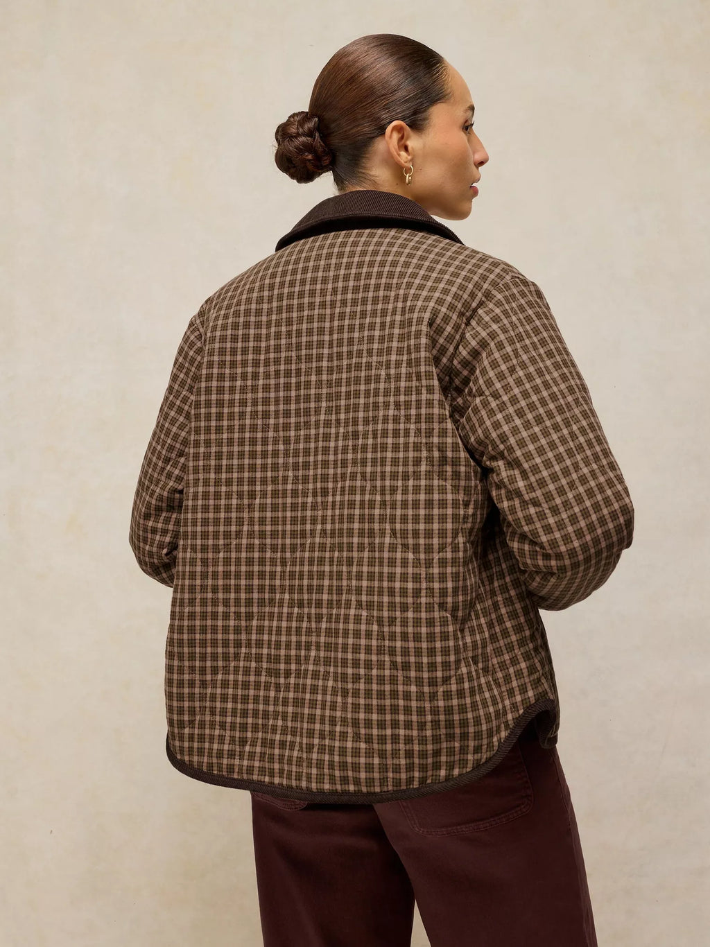 A woman shown from the back wearing a brown quilted plaid jacket with dark trim, paired with dark brown pants, against a textured beige background.