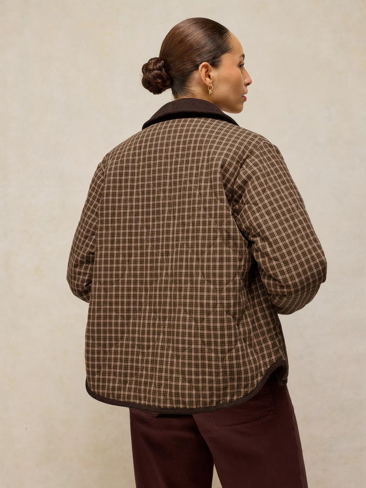 A woman shown from the back wearing a brown quilted plaid jacket with dark trim, paired with dark brown pants, against a textured beige background.