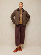A model stands against a neutral beige studio backdrop wearing a brown and black checkered jacket over a tan sweater, paired with dark plum cropped trousers and rust-colored sneakers.