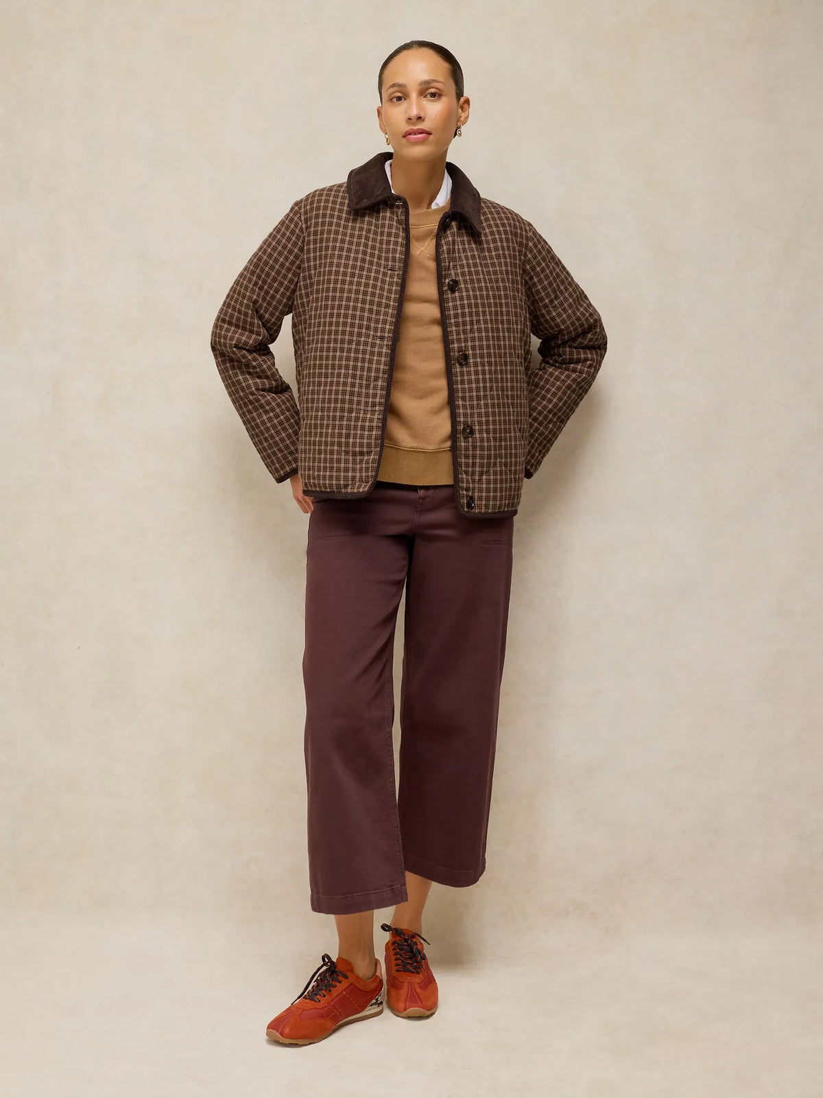 A model stands against a neutral beige studio backdrop wearing a brown and black checkered jacket over a tan sweater, paired with dark plum cropped trousers and rust-colored sneakers.
