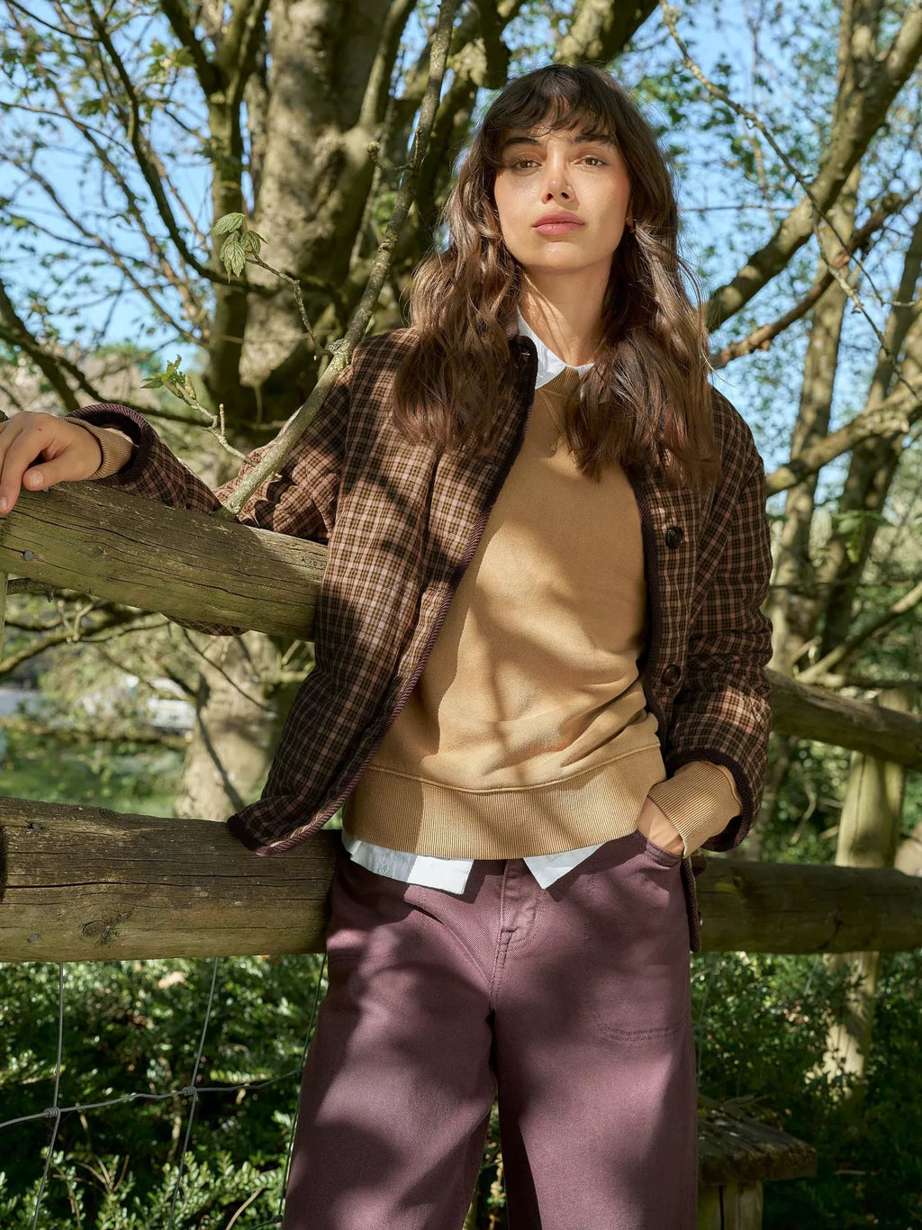 A woman stands outdoors leaning on a wooden fence, wearing a brown plaid overshirt over a tan sweater, a white collared shirt, and mauve-colored trousers. Trees and foliage fill the background.