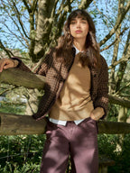 A woman stands outdoors leaning on a wooden fence, wearing a brown plaid overshirt over a tan sweater, a white collared shirt, and mauve-colored trousers. Trees and foliage fill the background.