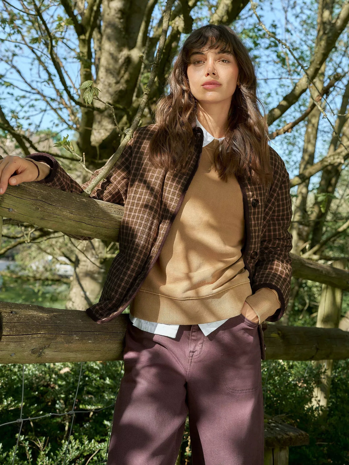 A woman stands outdoors leaning on a wooden fence, wearing a brown plaid overshirt over a tan sweater, a white collared shirt, and mauve-colored trousers. Trees and foliage fill the background.