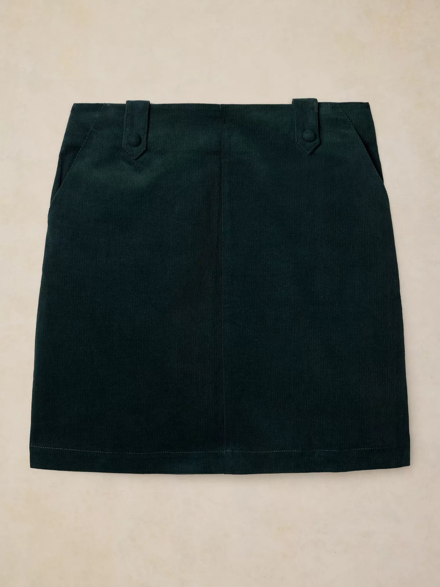 A dark forest green mini skirt with belt loops and side pockets, laid flat on a light beige textured background.