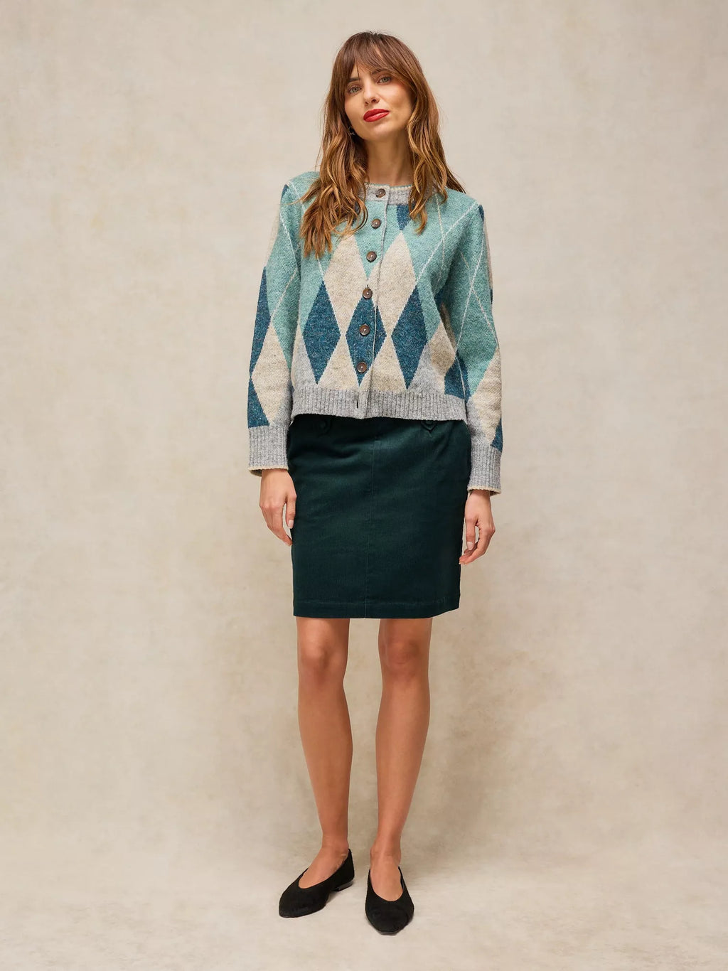 A woman stands against a textured beige studio backdrop wearing a patterned button-front cardigan and a dark green pencil skirt with black flats.
