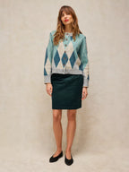 A woman stands against a textured beige studio backdrop wearing a patterned button-front cardigan and a dark green pencil skirt with black flats.