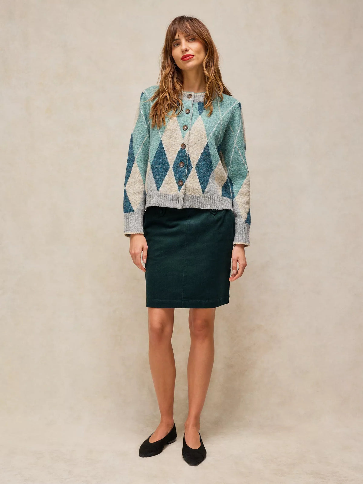 A woman stands against a textured beige studio backdrop wearing a patterned button-front cardigan and a dark green pencil skirt with black flats.