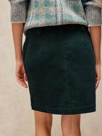 A dark forest green mini skirt with a double button closure at the waist, worn by a model styled with a gray and teal plaid sweater.