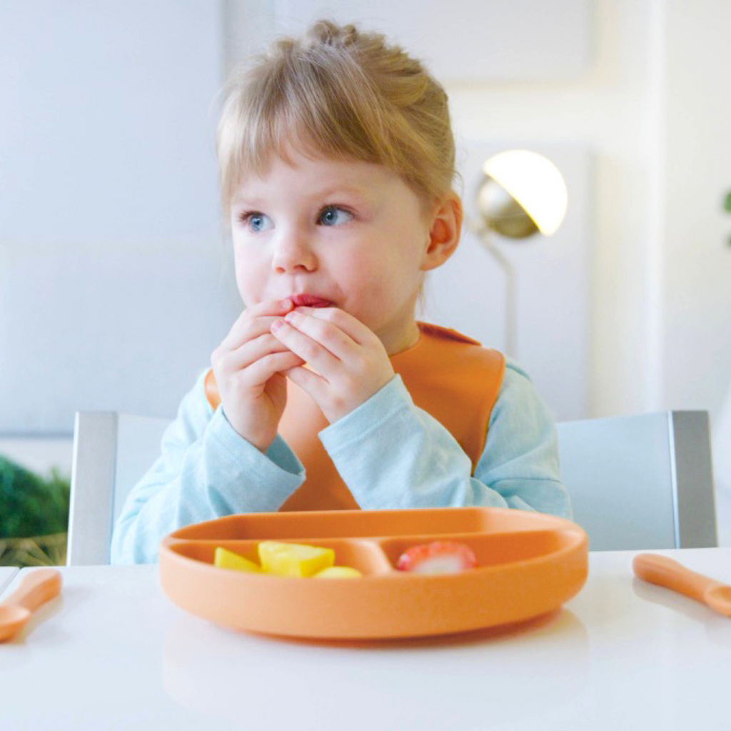 Child sitting at a table with an orange plate and bib, eating.