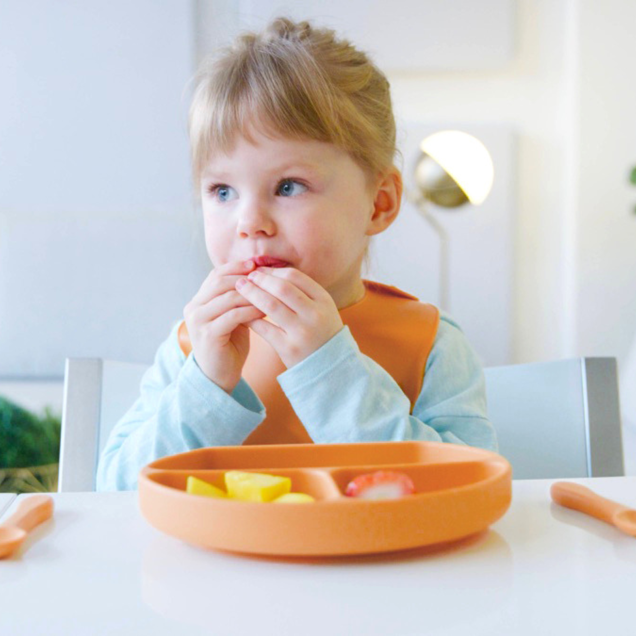 Child sitting at a table with an orange plate and bib, eating.