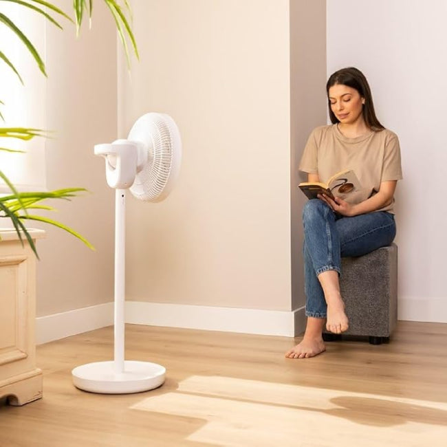 Woman reading a book next to a Cecotec EnergySilence 2600 Sunflower Fan in White - full view in a room with beige walls and wooden flooring.