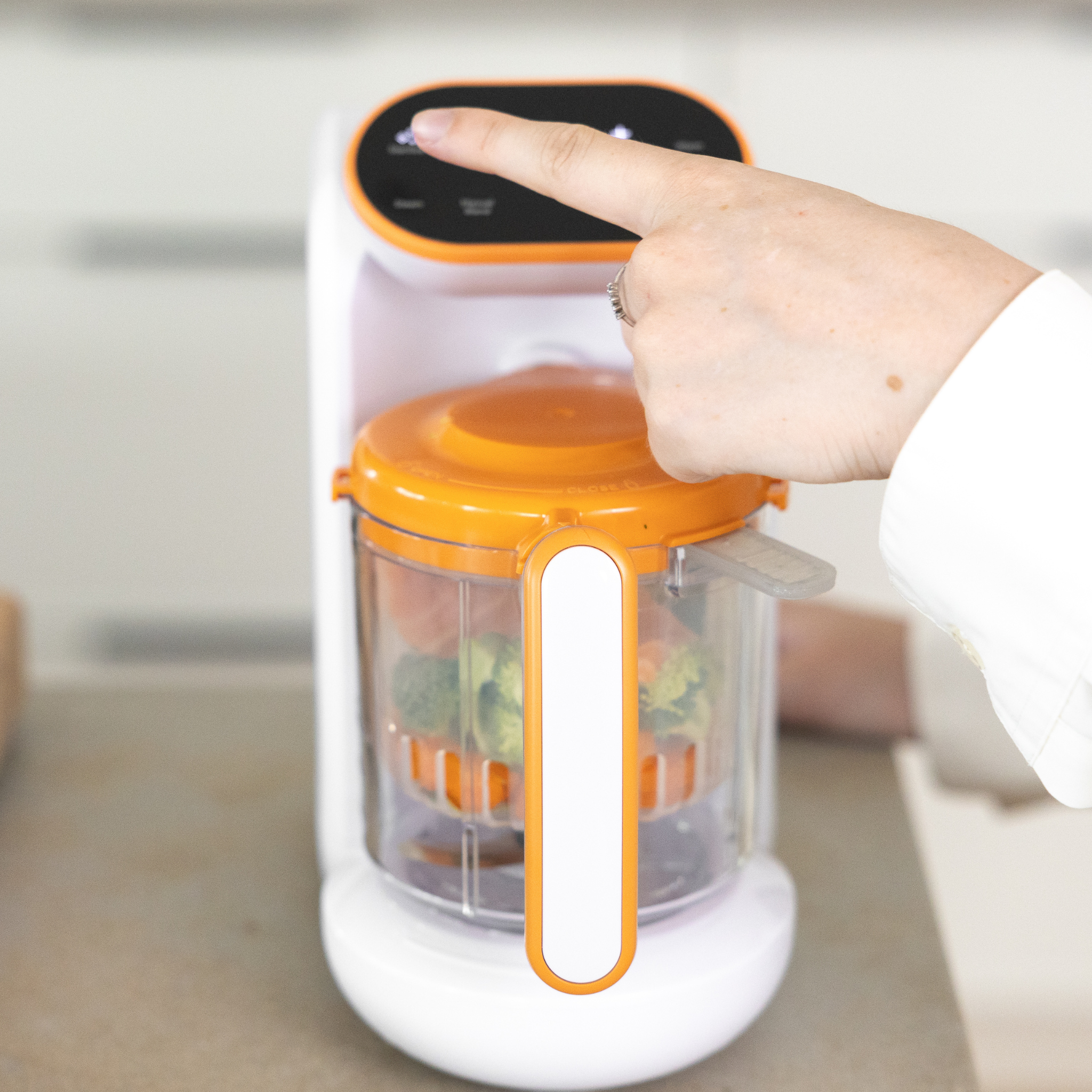 Hand pressing a button on a white and orange kitchen appliance with a transparent container holding fruits.