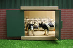 Model of a barn with toy cows inside, set against a brick wall and green grass background.
