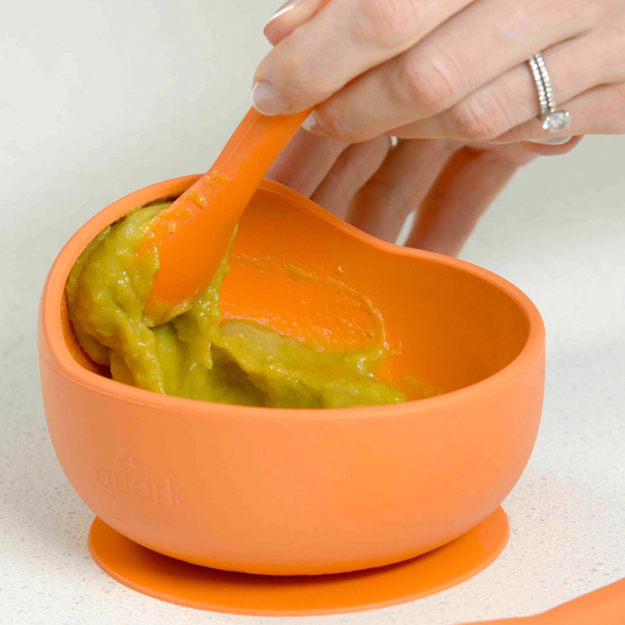 Orange baby bowl with green puree and orange spoon on a white background