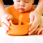 Child wearing an orange bib with a bowl and spoon, held by adult hands.
