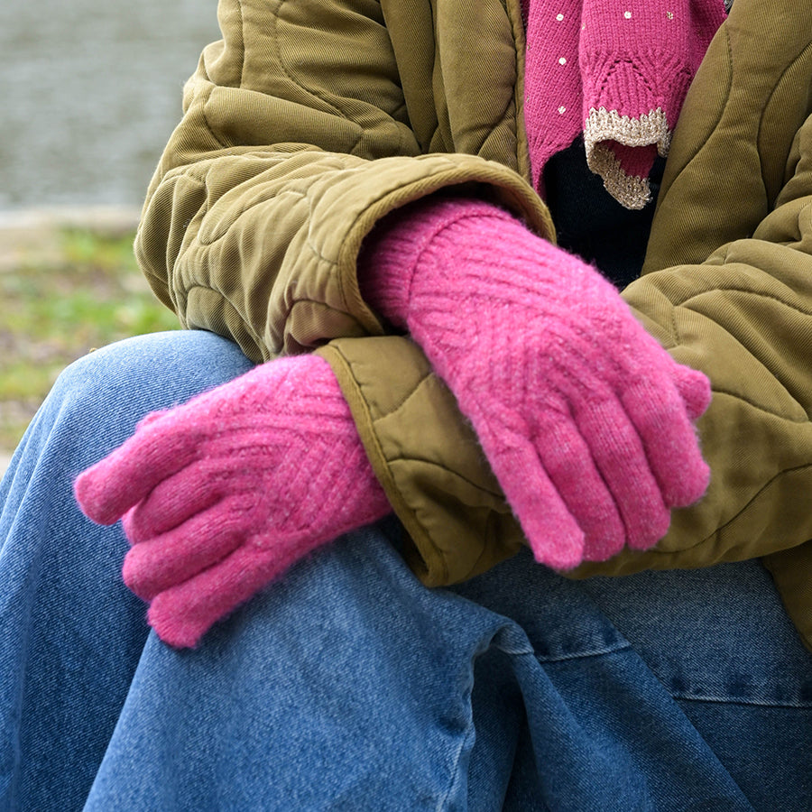 A close-up of a person sitting outdoors wearing bright pink knit gloves, an olive green quilted jacket, a pink knit sweater, and blue jeans.