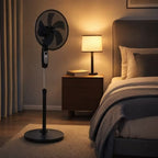 Crystal Home Air Comfort 40cm Pedestal Fan in Black - full view on a carpeted floor next to a bed in a warmly lit bedroom. A wooden nightstand with a lit table lamp and a book sits between the fan and the bed.