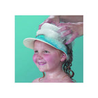 A child wearing a blue shampoo eye shield cap with a turquoise band, having their hair washed with soap while an adult's hands wash the hair, set against a solid turquoise background.