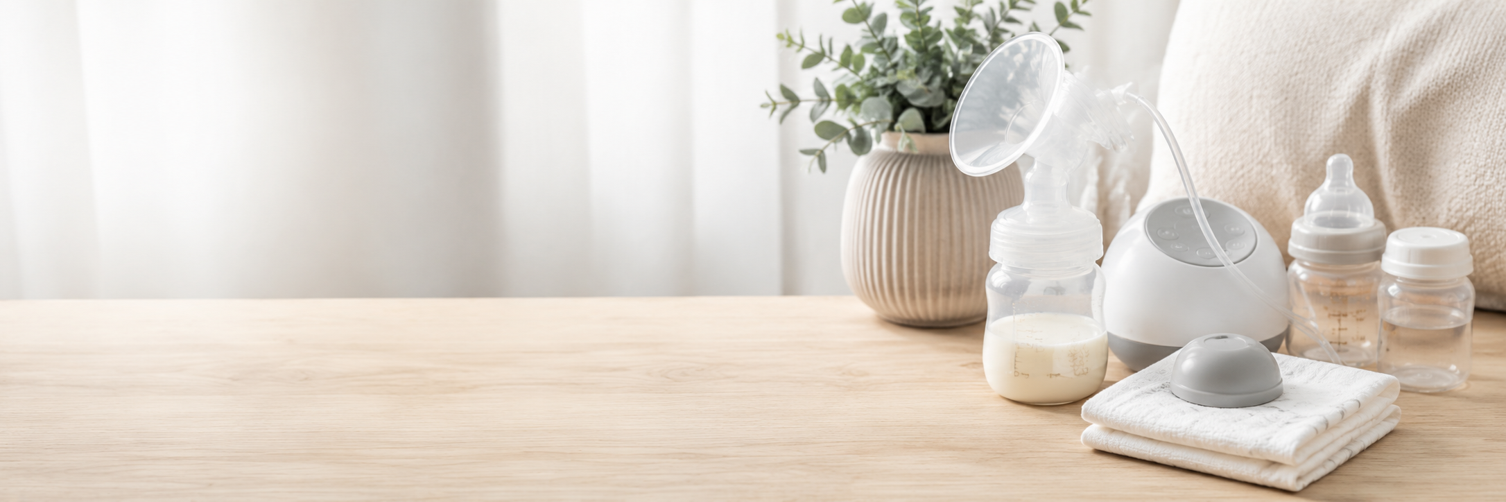 Breastfeeding and pumping supplies arranged on a light wood table in a softly lit room, including a breast pump, bottles, a folded cloth, and a small vase with greenery.