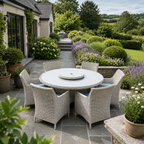 A round outdoor dining set with a light gray stone-look table and six woven wicker chairs with blue cushions, placed on a stone patio in a landscaped garden courtyard.