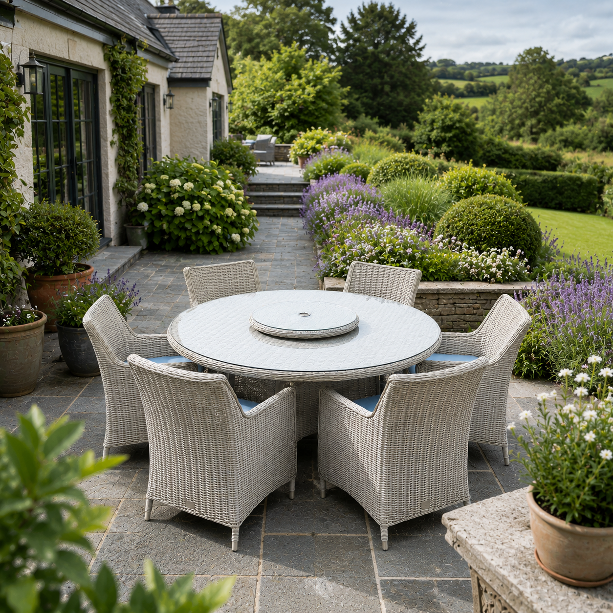 A round outdoor dining set with a light gray stone-look table and six woven wicker chairs with blue cushions, placed on a stone patio in a landscaped garden courtyard.