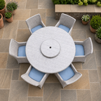 A 7-piece outdoor patio dining set featuring a round light gray woven table with a built-in umbrella hole and six matching gray wicker chairs with light blue cushions, arranged on a stone patio.