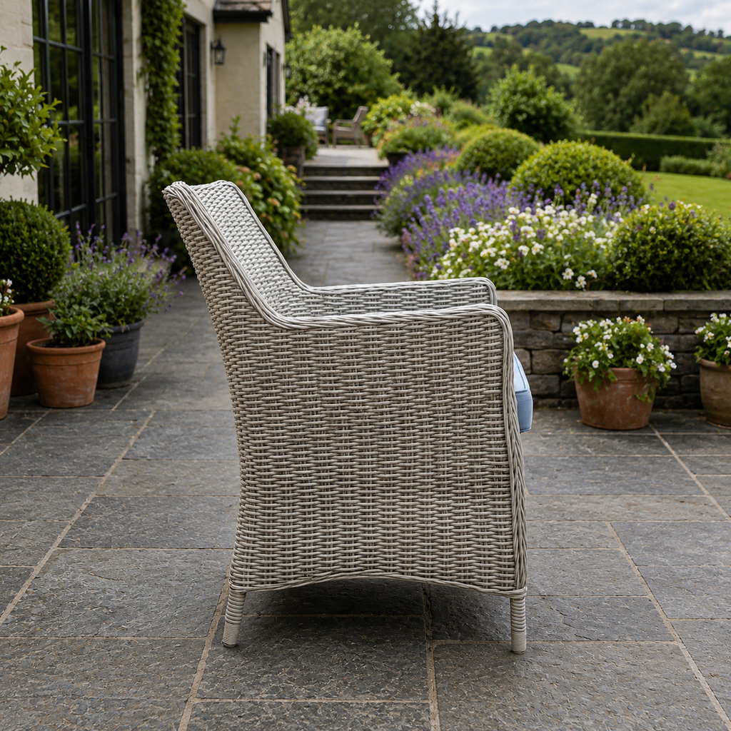 A light gray woven wicker outdoor armchair is shown from a side-rear angle on a stone patio in a landscaped garden setting.
