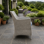 A light gray woven wicker outdoor armchair is shown from a side-rear angle on a stone patio in a landscaped garden setting.