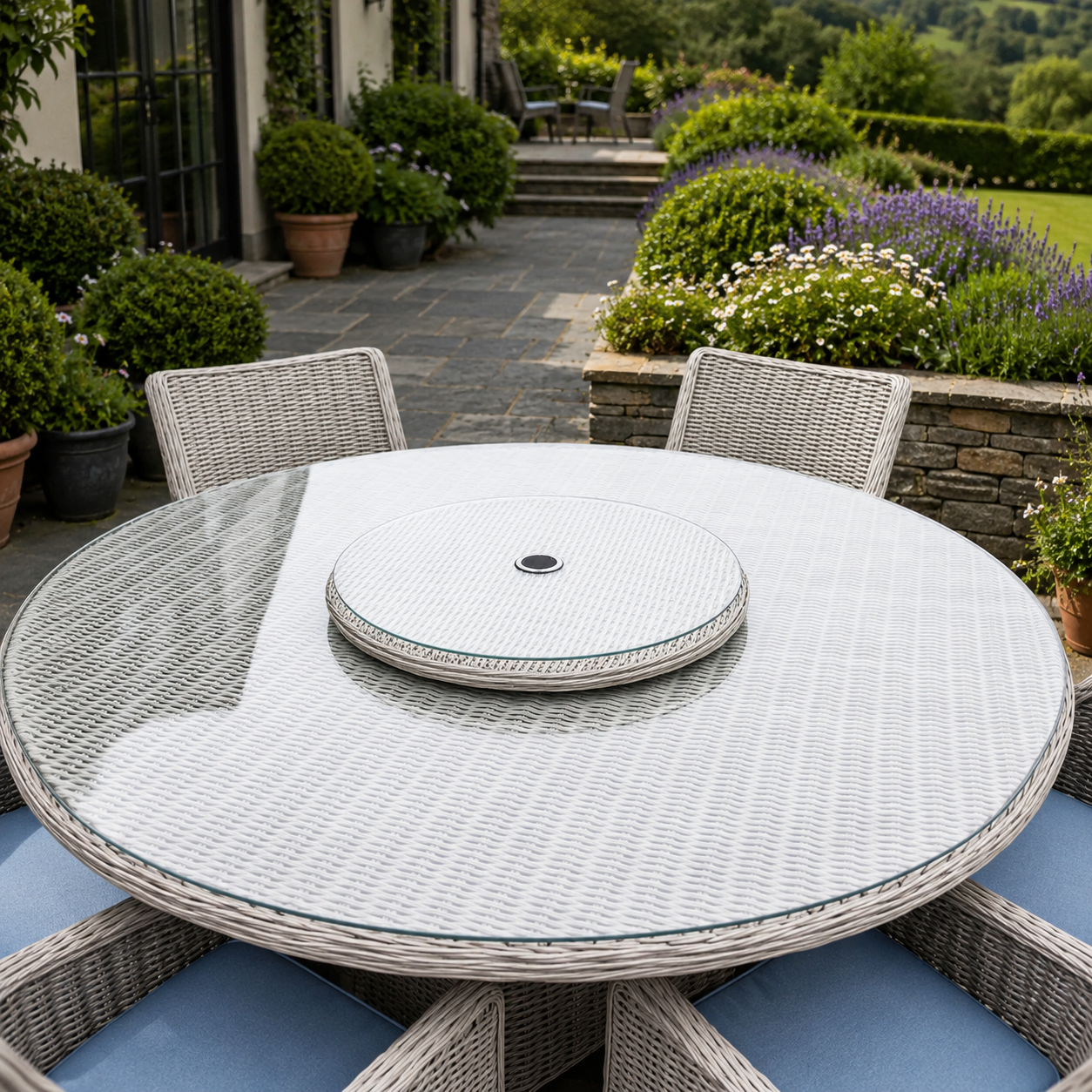 Round outdoor dining table set with a woven wicker-style frame, clear glass tabletop, matching chair backs, and a round center insert, shown in a landscaped patio garden.