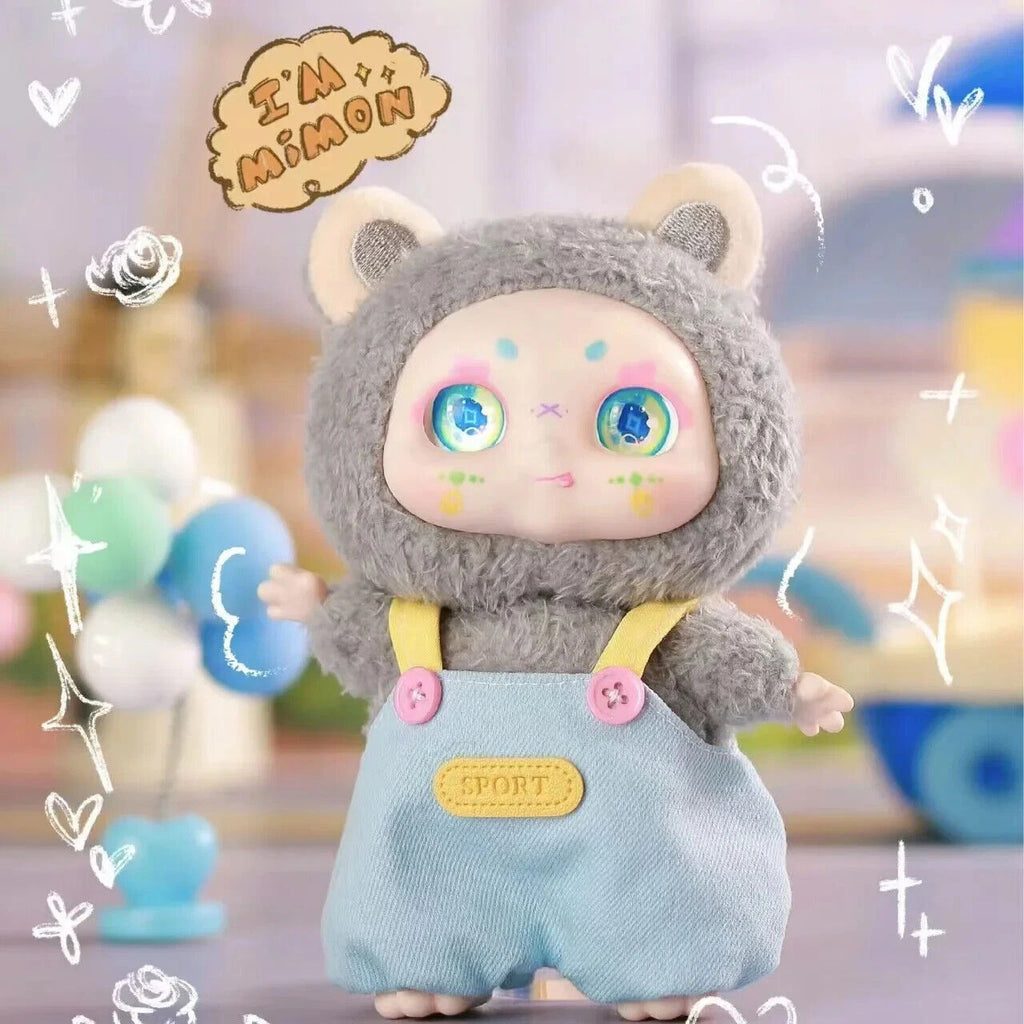 Toy figure with cat-like design wearing a gray hood and light blue overalls with pink buttons, standing against a colorful blurred background.