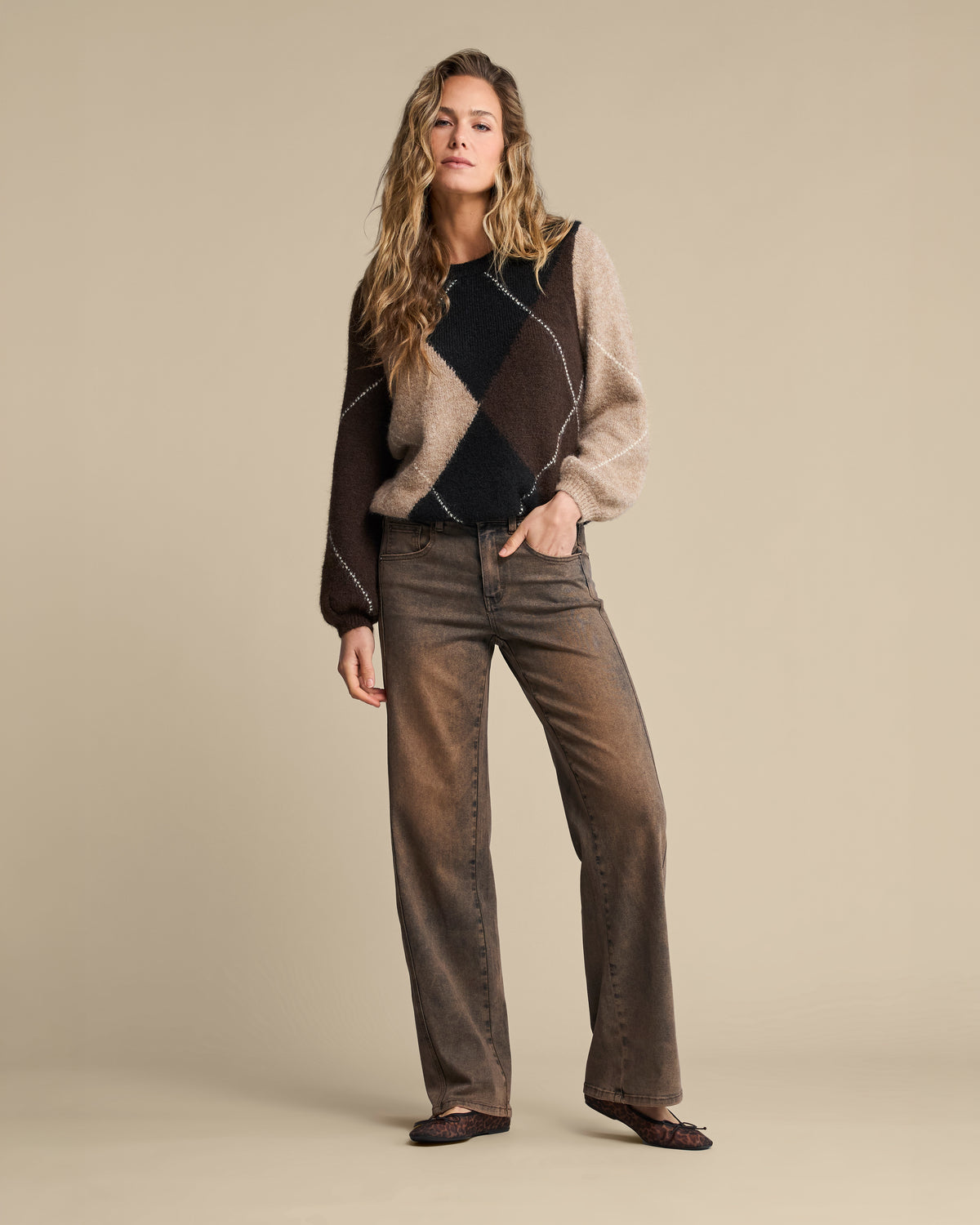 A woman modeling a brown and beige argyle sweater with dark brown wide-leg jeans and dark brown flats, standing against a solid beige studio background.