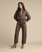 A woman modeling a brown leopard-print long-sleeve blouse and brown straight-leg pants with taupe heeled ankle boots, standing against a solid beige studio background.