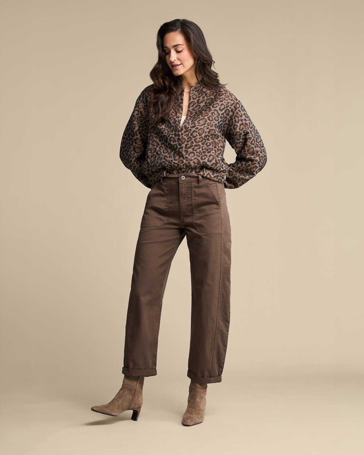 A woman modeling a brown leopard-print long-sleeve blouse and brown straight-leg pants with taupe heeled ankle boots, standing against a solid beige studio background.