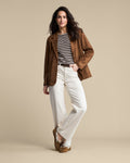 A woman models a brown suede blazer over a black and white striped t-shirt, paired with off-white wide-leg jeans and brown suede mules, set against a solid beige studio background.