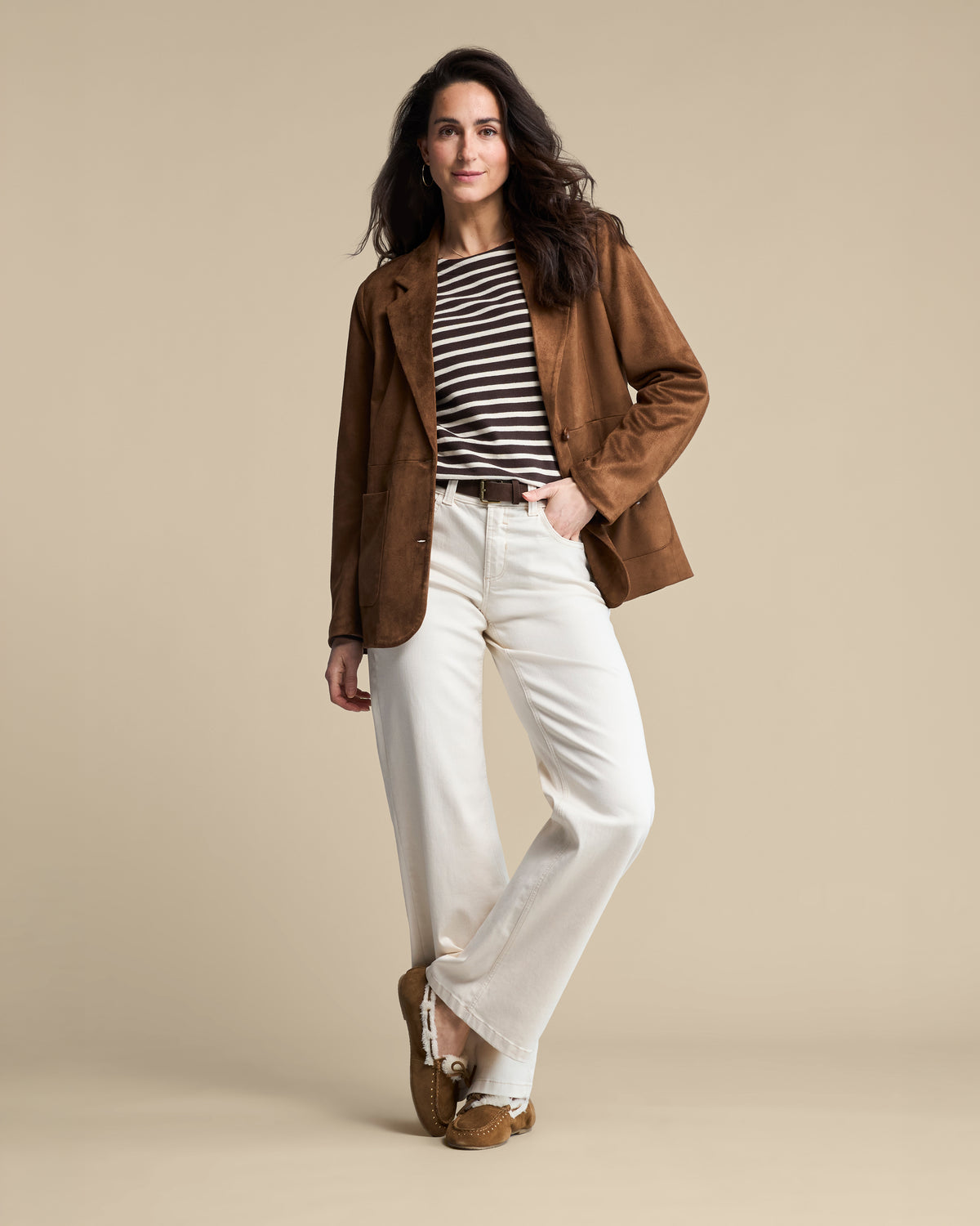 A woman models a brown suede blazer over a black and white striped t-shirt, paired with off-white wide-leg jeans and brown suede mules, set against a solid beige studio background.