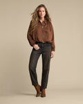 A woman modeling a brown satin zip-up bomber jacket, dark charcoal gray jeans, and brown leather boots against a solid beige studio background.