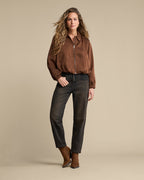 A woman modeling a brown satin zip-up bomber jacket, dark charcoal gray jeans, and brown leather boots against a solid beige studio background.