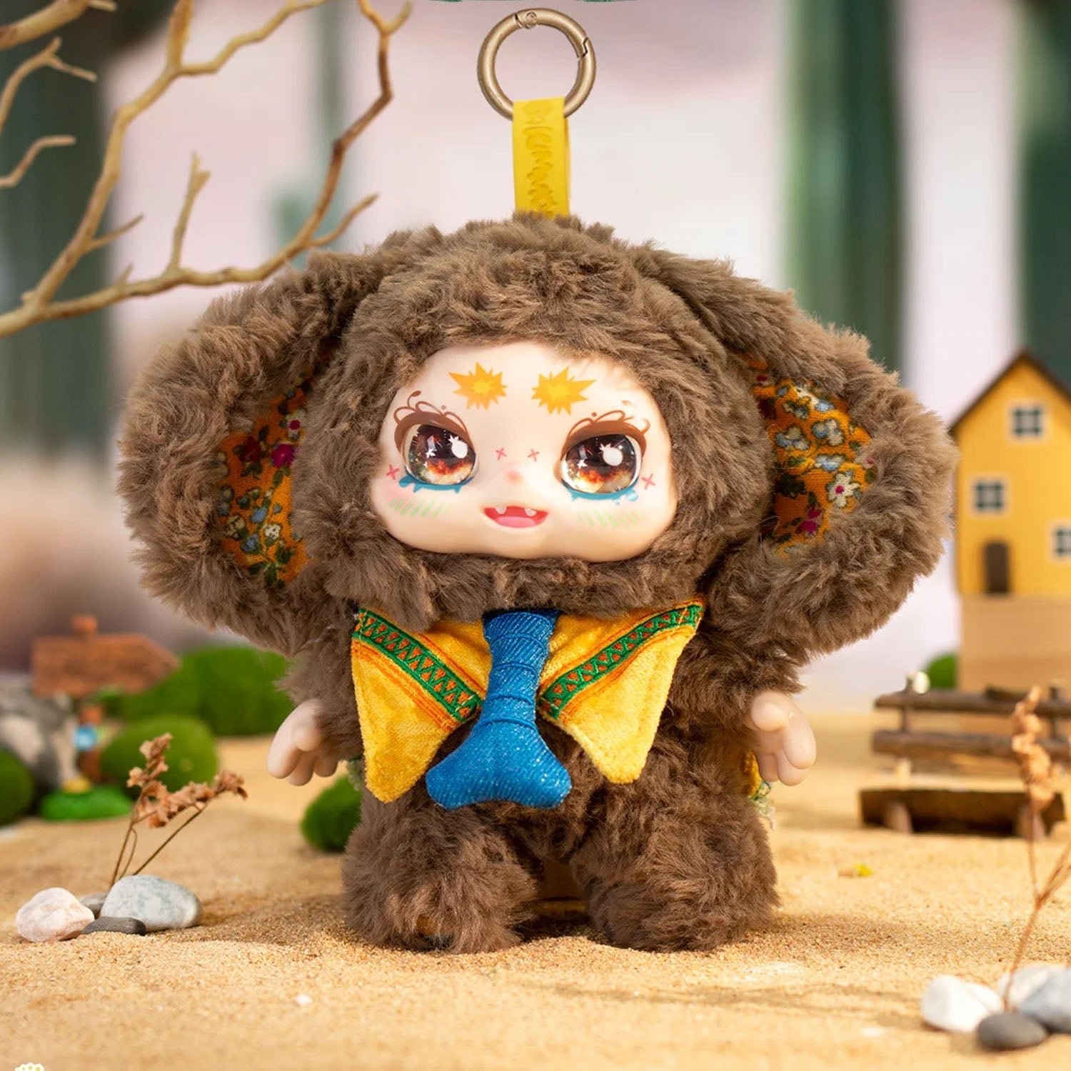 Plush toy with a bear-like design wearing a yellow outfit with a blue bow tie, standing on a sandy surface with a blurred natural background.