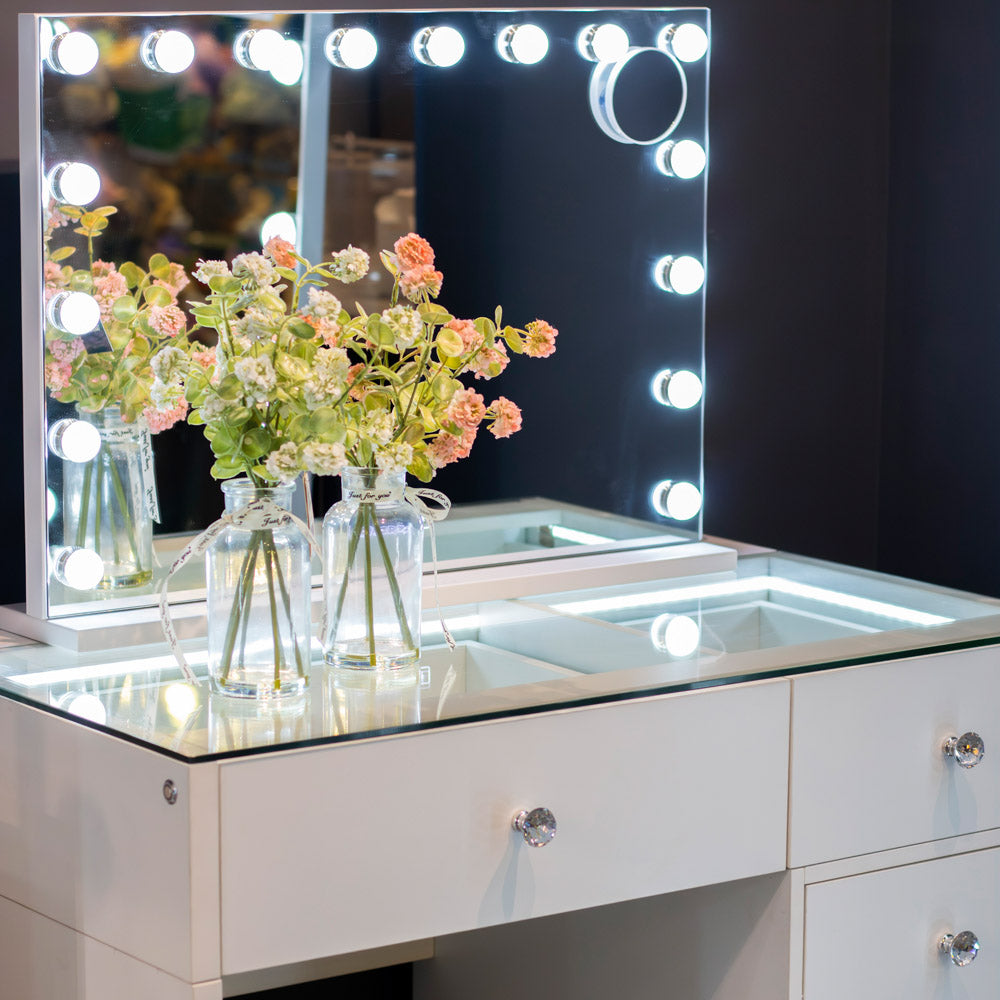 Makeup vanity with a mirror with lights and flowers on the counter.
