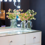 Two clear glass jars filled with pink and white faux flowers sit on a white vanity table with a glass top, in front of a lit mirror.