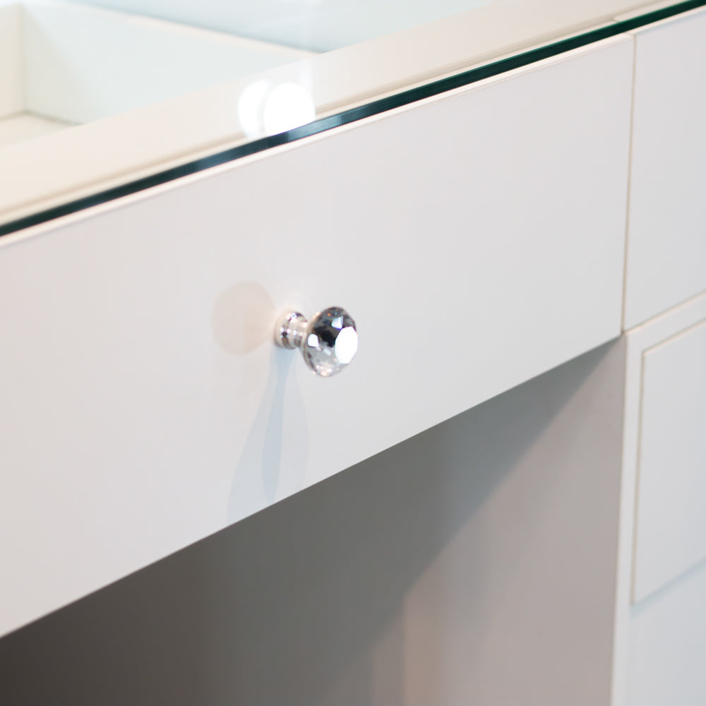 A close-up of a white wooden vanity drawer featuring a faceted clear crystal knob, with a glass top edge visible above.