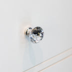 A clear faceted crystal cabinet knob with a polished silver-toned metal base, mounted on a white wooden surface.