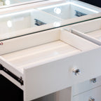Close-up of a white vanity or dresser drawer with a glass top and illuminated mirror, showing the open drawer interior and silver knobs.
