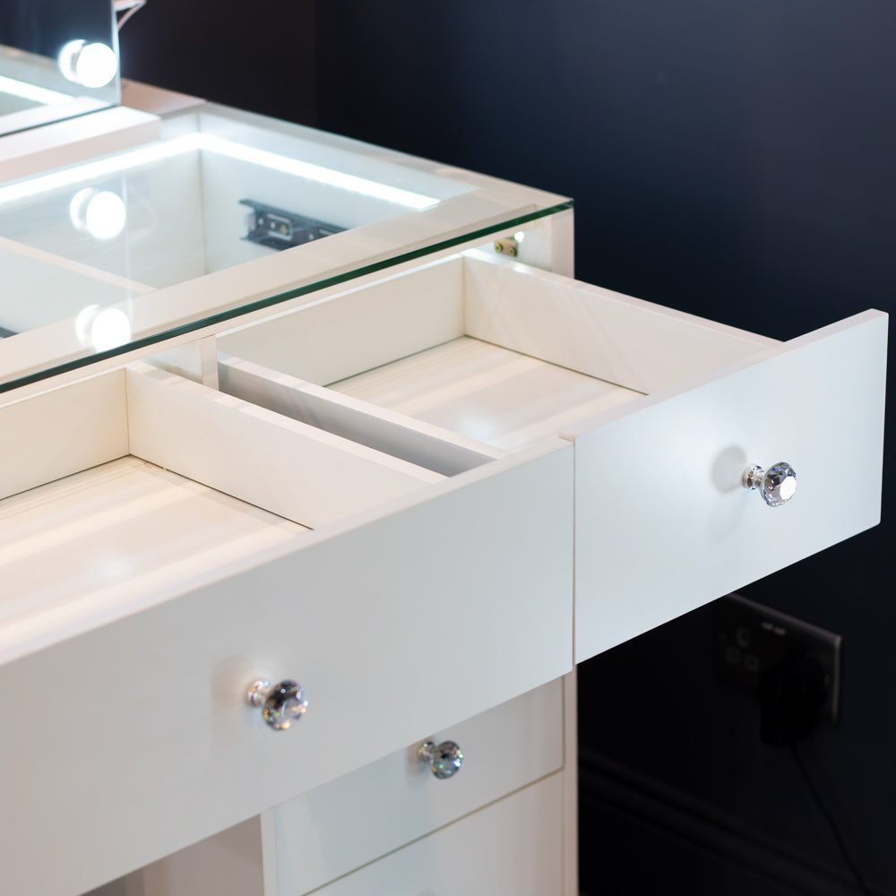 A close-up of a white vanity or desk with an open drawer showing interior compartments, crystal-style drawer knobs, a glass top edge, and a dark wall in the background.