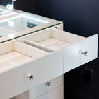 A close-up of a white vanity or desk with an open drawer showing interior compartments, crystal-style drawer knobs, a glass top edge, and a dark wall in the background.