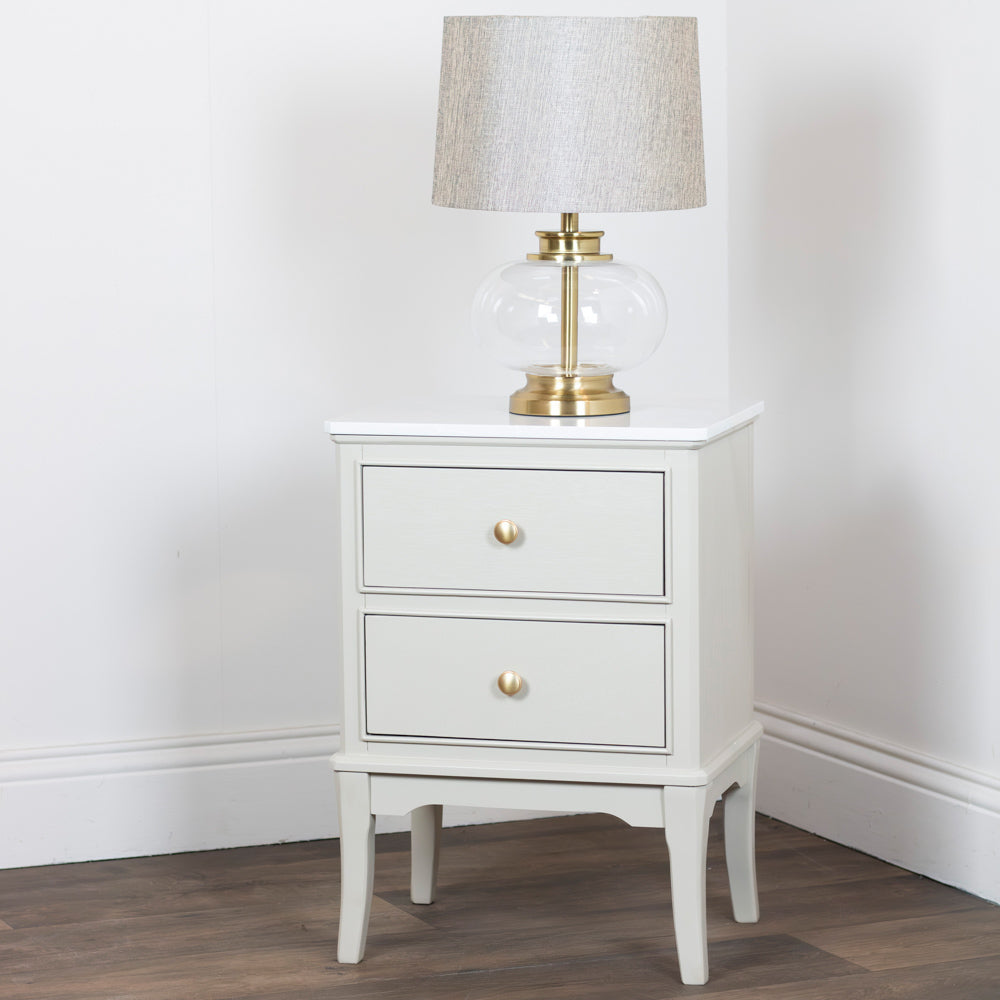 Monroe 2 Drawer Accent Table in Stone with two drawers and a lamp on a wooden floor against a white wall.