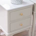 A close-up of a two-drawer Monroe 2  Drawer Accent Table in Stonewith a white marble-look top, a warm off-white painted body, and round gold knobs, shown in a styled interior setting.