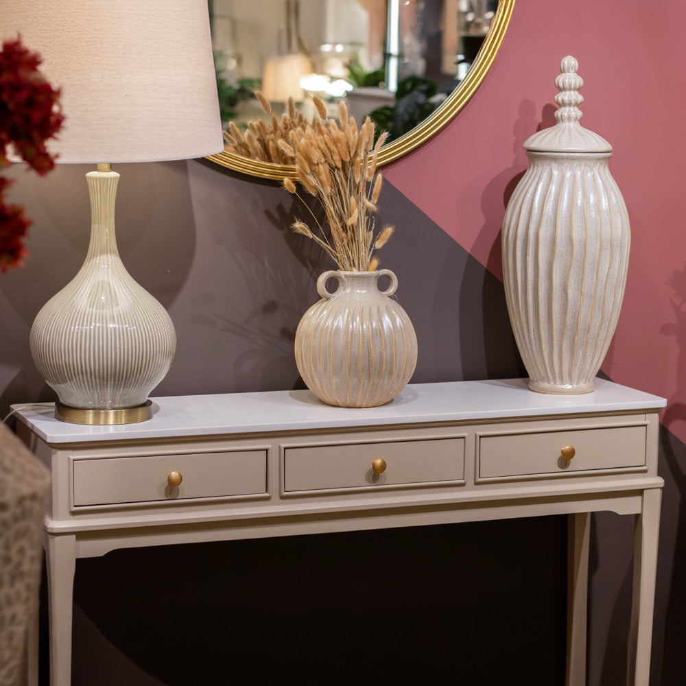 A styled interior scene showing a console table with a white marble top, three drawers with gold knobs, and decorative ceramic vases and a lamp against a pink and gray wall with a gold-framed mirror.