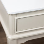 Close-up of a white wooden furniture piece, showing the corner of the top and a drawer front with decorative molding.
